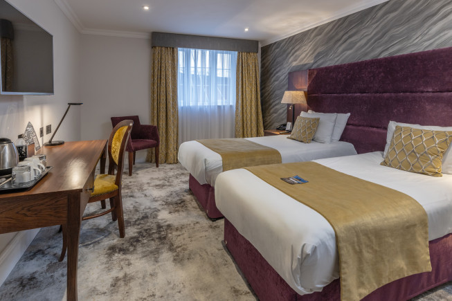 A hotel room with two twin beds, a desk and chair, a lamp, and a window with curtains.