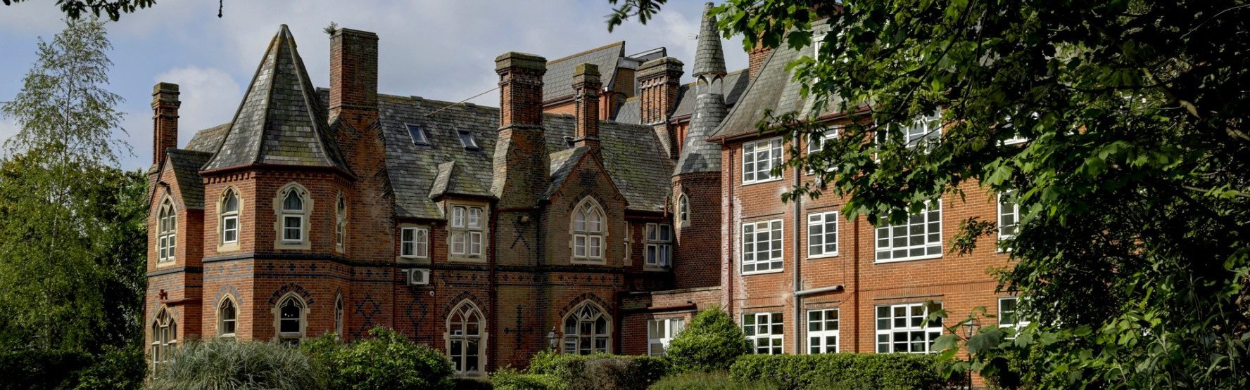 A large historic brick building with steep slate roofs, pointed turrets, and many windows, partly framed by trees and shrubs.