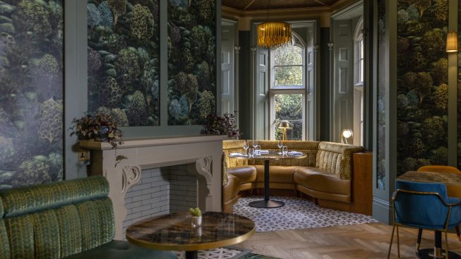 Interior of a decorated lounge with patterned wallpaper, a fireplace, booth seating by a window, and small tables.
