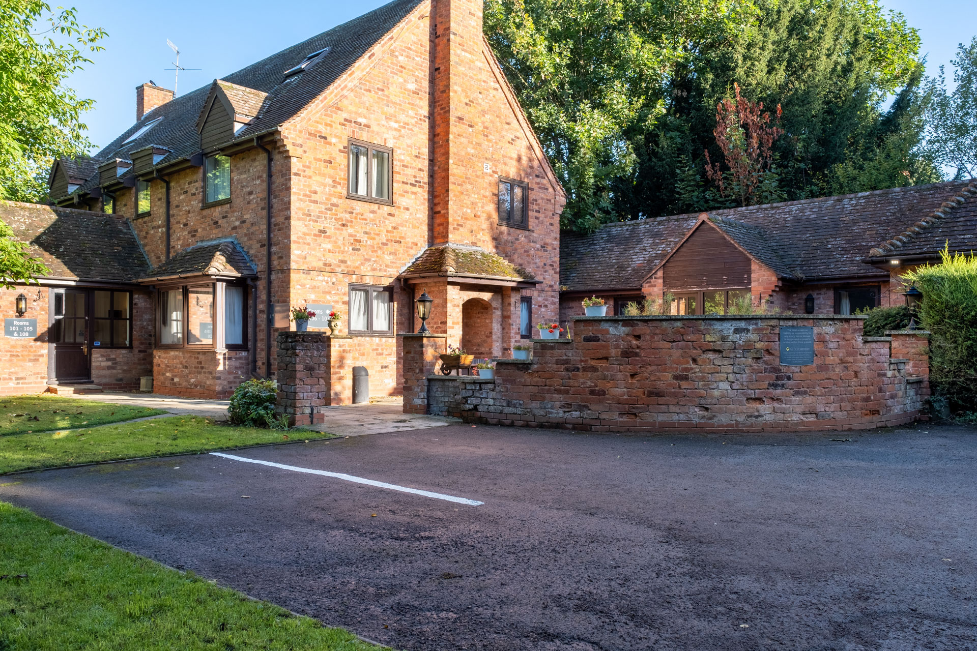 A large brick house with steep roofs, small windows, and a walled courtyard beside a driveway and trees.