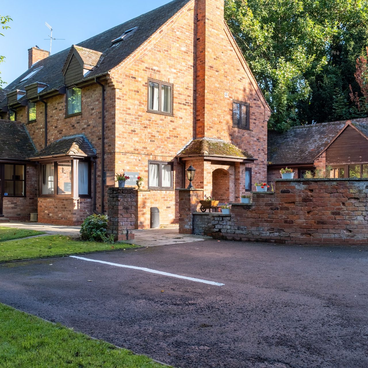 A large brick house with steep roofs, small windows, and a walled courtyard beside a driveway and trees.