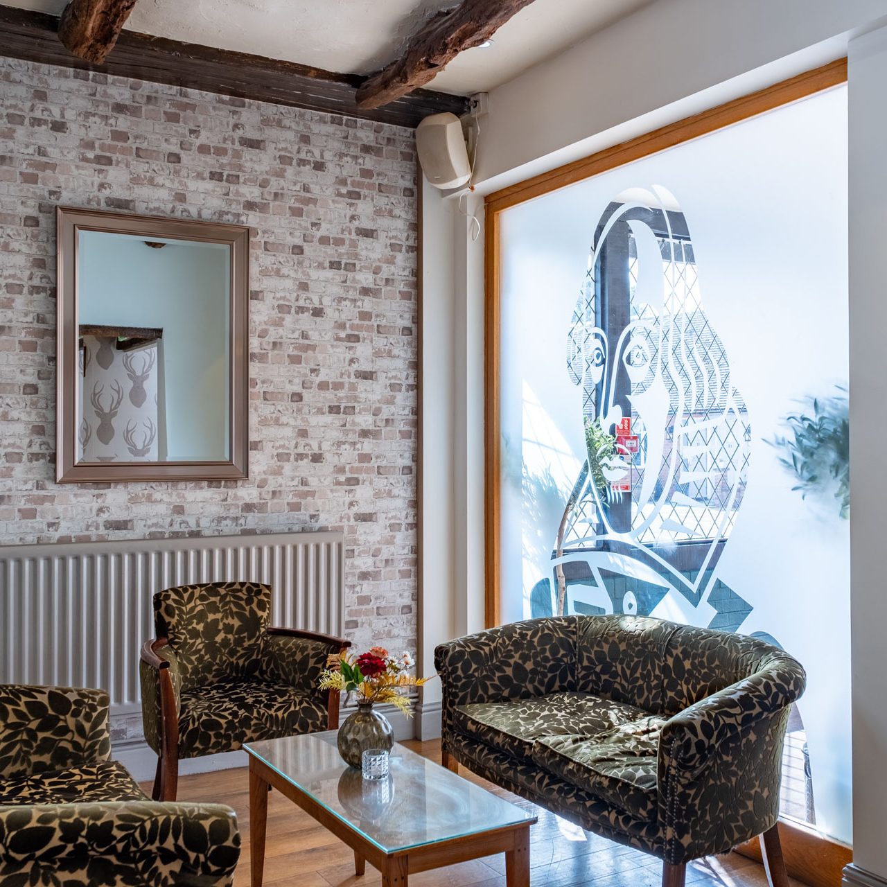 A small lounge with patterned armchairs and sofa around a glass coffee table, a brick accent wall, framed wall art, and a large window with-