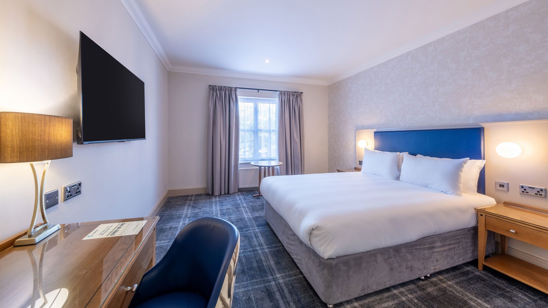 A modern hotel bedroom with a large bed, wall-mounted TV, desk, lamp, and window with curtains.