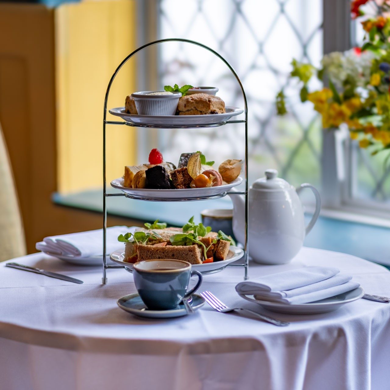A three-tier stand with sandwiches, scones, and pastries on a table set with cups, plates, and a white teapot beside a window.