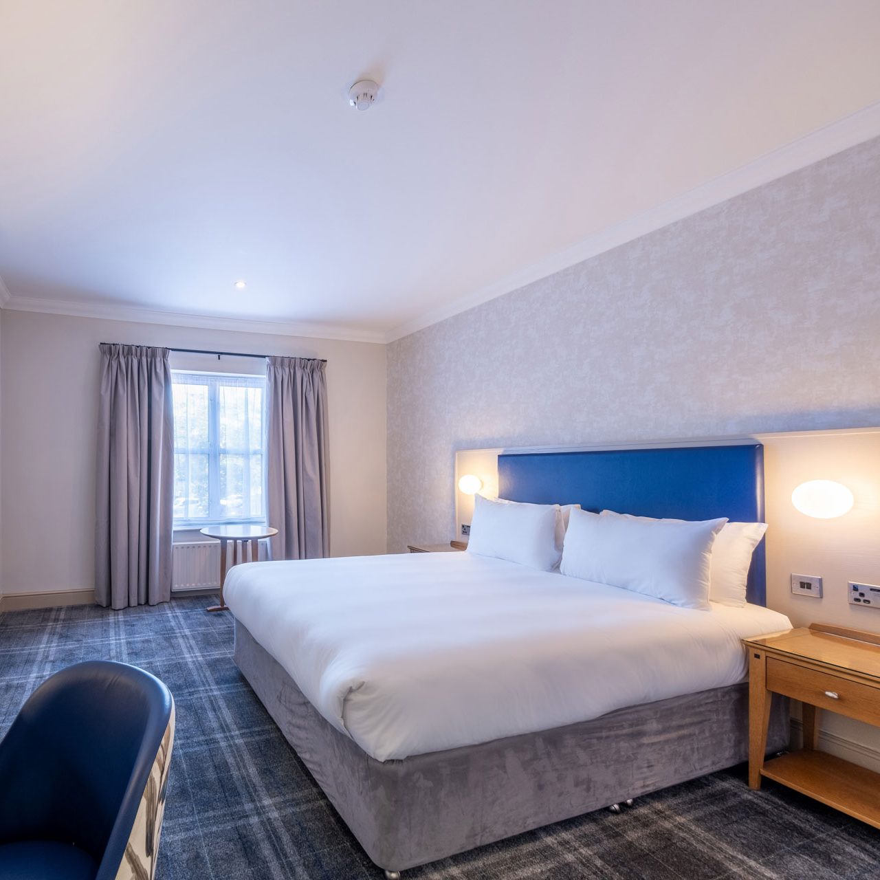 A modern hotel bedroom with a large bed, wall-mounted TV, desk, lamp, and window with curtains.