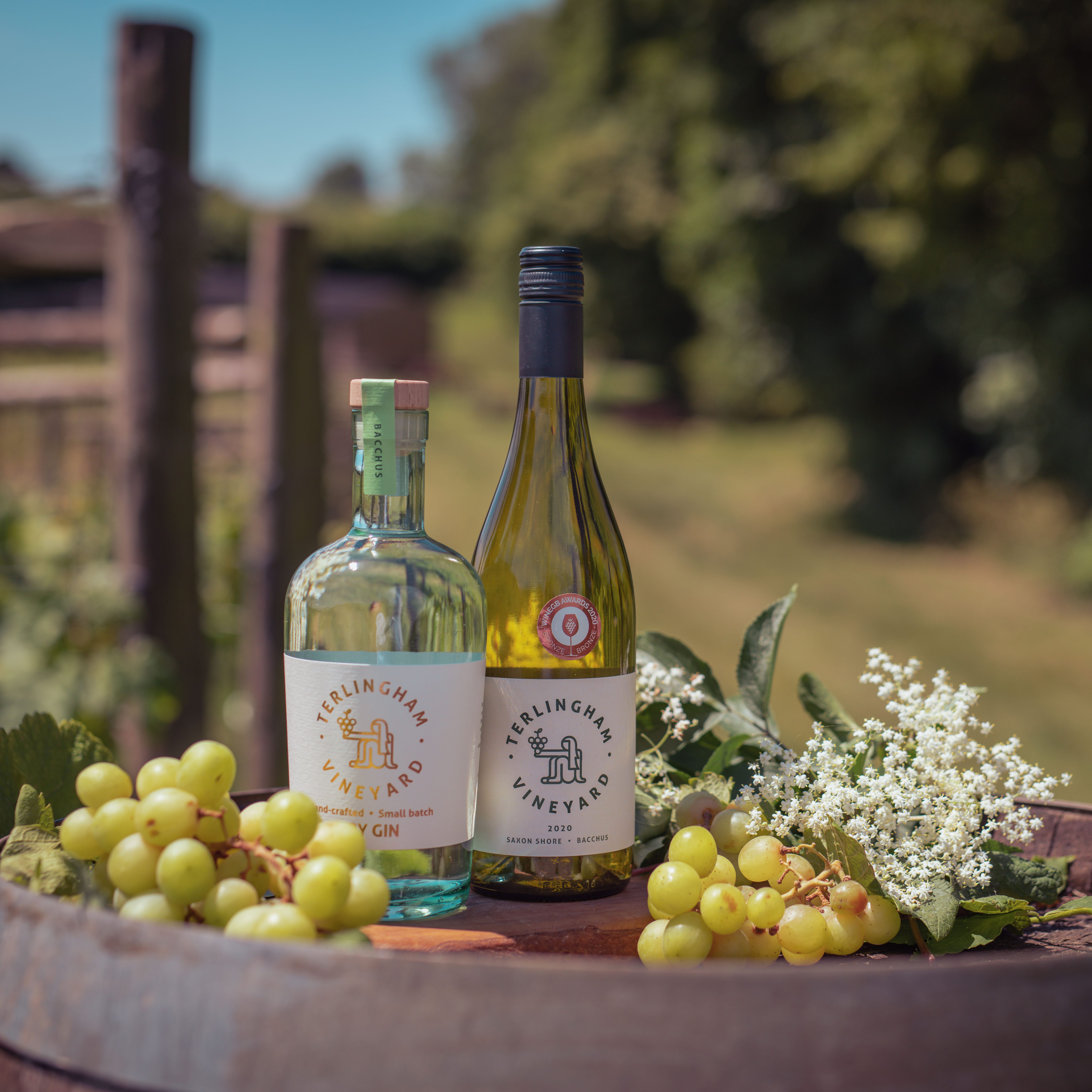Two wine bottles standing on a barrel outdoors, surrounded by green grapes and white flowers with a blurred vineyard background.