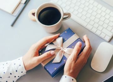 Hands holding a blue gift box with a ribbon beside a coffee cup