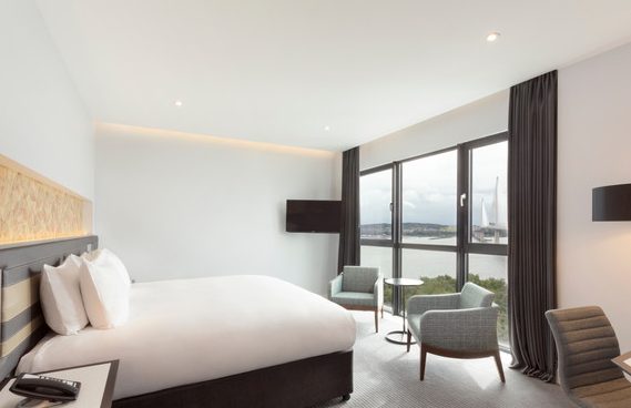 Bright hotel room featuring a large bed and windows overlooking water.