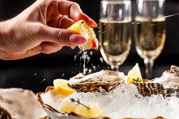 A hand squeezing lemon over a plate of oysters with champagne glasses in the background