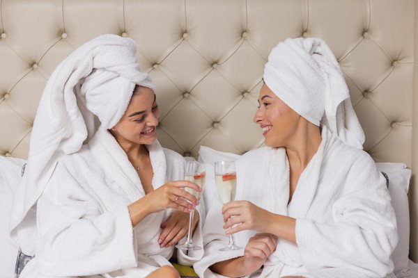 Two women in white robes and towels toast with champagne glasses